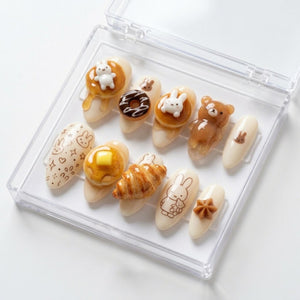 A set of false nails in a clear plastic case. The nails feature various designs, including a bear, a bunny, a croissant, a donut, and pancakes with syrup and butter. Some nails have bunny illustrations.