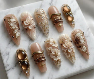 A collection of ten almond-shaped artificial nails. The nails feature various designs, including floral embellishments, gold accents, and jewel-like adornments. Colors include nude, gold, brown, and white. Some nails have a butterfly wing design.