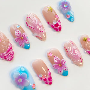 A collection of artificial nails with various designs. Some nails feature pink floral accents, while others have abstract patterns in pink and white. Several nails have a nude base with colorful embellishments and a turquoise or blue base.