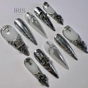 A collection of ten long, pointed artificial nails. The nails feature a combination of silver metallic and white designs, including crosses, floral accents, and intricate patterns. The brand name "IRIS NAILART" is visible in the upper left corner.