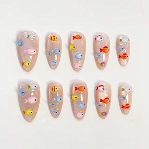 Set of ten artificial nails with a nude base, each adorned with colorful, cartoon fish designs. The fish are in shades of yellow, orange, pink, blue, and silver, creating a playful, aquatic theme. The nails are oval-shaped.