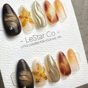 A set of ten almond-shaped press-on nail tips displayed on a white textured card labeled LeStar Co. The designs include dark brown, translucent amber, gold metallic wavy textures, and 3D floral accent