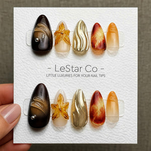 A set of ten press-on nails arranged on a white display card labeled LeStar Co - LITTLE LUXURIES FOR YOUR NAIL TIPS. The nails feature almond shapes with varied designs including dark brown, amber flo
