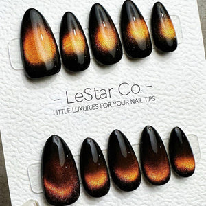A set of ten almond-shaped press-on nails with a glossy black base and a shimmering orange cat-eye magnetic effect in the center. The nails are displayed on a white card labeled LeStar Co - LITTLE LUX