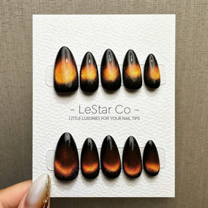 A set of ten press-on nails arranged on a white display card with the text LeStar Co - LITTLE LUXURIES FOR YOUR NAIL TIPS. Each nail features a glossy black base with a shimmering, cat-eye style orang