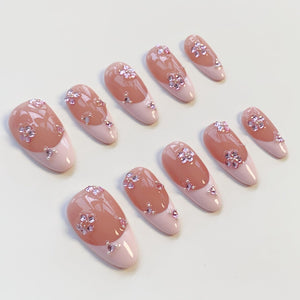 Set of ten artificial nails with a light pink base and a peach-colored top. Each nail features a floral design made of small, sparkling rhinestones. The nails have a rounded shape.