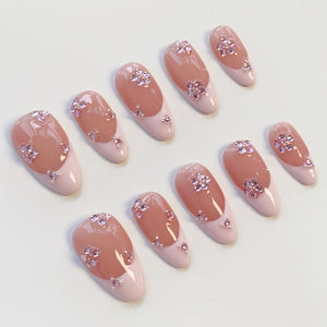 A set of ten artificial nails with a light pink base and a French tip design. Each nail features a floral embellishment made of small, sparkling crystals. The nails have an oval shape and a glossy finish, perfect for a special occasion.