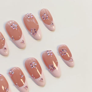 Oval-shaped artificial nails with a light pink base and a darker pink top. Each nail features a flower design made of small, pink rhinestones. Additional rhinestones are placed near the base of each nail.