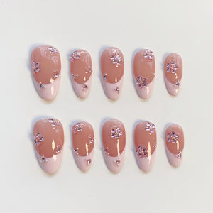 Set of ten artificial nails. Each nail features a light pink base with a darker pink tip. The nails are adorned with small, sparkling, pink floral embellishments. The nails are oval-shaped.