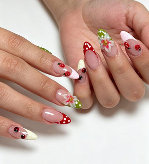 Almond-shaped nails featuring a French manicure base with various decorative elements including red strawberries, white polka dots, green accents, and hand-painted white flowers with yellow centers on a clear, glossy nail polish finish.