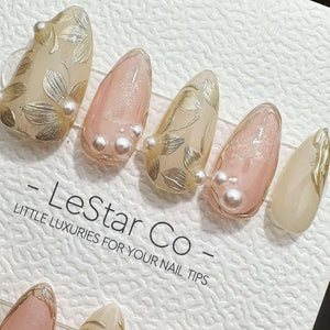 A set of almond-shaped press-on nails featuring a mix of nude and soft pink tones, decorated with metallic gold leaf floral patterns and small white pearl accents. The text LeStar Co - LITTLE LUXURIES