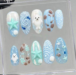 A set of ten press-on nails featuring an ocean theme with light blue, white, and mint green colors. The designs include raised 3D elements such as a white seal, a mint green starfish, small seashells, swirling water patterns, and various fish.