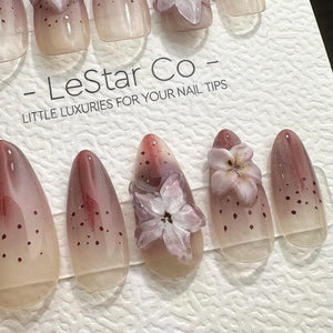 A set of almond-shaped false nails with a gradient design, transitioning from a nude base to a deep red at the tips. Some nails feature small red dots and delicate, three-dimensional flower embellishm