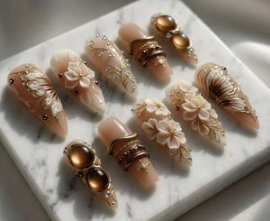 A collection of ten almond-shaped artificial nails. Each nail features intricate designs, including floral accents, gold filigree, and embedded gemstones. The nails are primarily nude with gold, brown, and white details, creating an elegant aesthetic