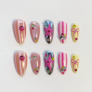 A set of ten artificial nails in various designs. Some nails feature pink and white stripes, ladybug accents, or colorful rhinestones. Others have a silver chrome finish with a pink star, or a yellow base with a pink bow.