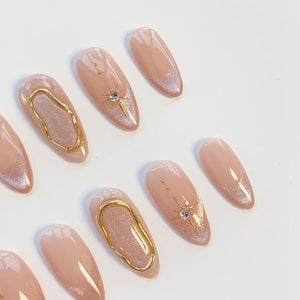 A set of almond-shaped press-on nails featuring a nude base with shimmering silver tips. Select nails are adorned with raised gold metallic swirls, while others feature a delicate gold starburst design with a small embedded clear crystal.