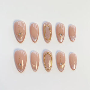 A set of ten almond-shaped press-on nails in a nude base color. The design features gold starburst accents with small embedded crystals, silver glitter tips, and a raised gold metallic organic oval frame on the middle fingernails.