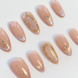 A set of almond-shaped press-on nails featuring a nude base with shimmering glitter tips. Several nails are adorned with gold metallic accents, including raised wavy lines and starburst designs centered with small clear rhinestones.