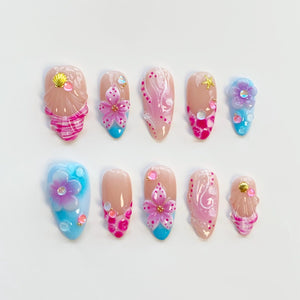A set of ten artificial nails with various designs. The nails feature a combination of pink, blue, and nude colors, with seashell, floral, and abstract patterns. Some nails have 3D embellishments.
