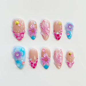 A set of ten artificial nails in various designs. The nails feature a nude base with pink, blue, and gold accents. Designs include flowers, seashells, and abstract patterns. The nails are oval-shaped.