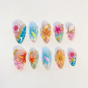 A set of ten press-on nails featuring a tropical theme with 3D embellishments. Designs include pink and orange hibiscus flowers, blue ocean waves, yellow and pink dolphins, starfish, seashells, and small gold beads on a nude base.