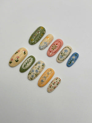 A set of ten oval-shaped press-on nails featuring various designs including floral patterns, checkered backgrounds, and landscape motifs in shades of sage green, pastel yellow, soft pink, cream, orange, and sky blue with fine details.