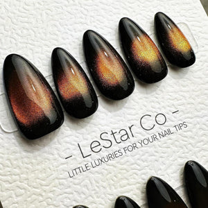 A set of almond-shaped press-on nails featuring a glossy black base with a shimmering, magnetic cat-eye effect in shades of orange and gold. The nails are displayed on a white card printed with the te