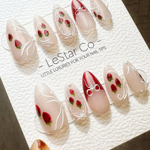 A set of almond-shaped press-on nails featuring a nude base with hand-painted red strawberries, white swirling line accents, and small white pearl embellishments. The display card reads LeStar Co. LIT