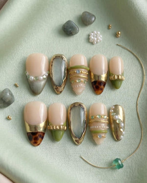 A set of ten almond-shaped press-on nails featuring a nude base with decorative accents including gold metallic bands, green and white beads, small rhinestones, and brown leopard print patterns, arranged in two rows on a light green fabric.