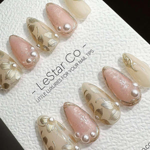 A set of almond-shaped press-on nails featuring a mix of soft pink and cream colors, adorned with gold leaf floral patterns and small white pearls. The text LeStar Co. LITTLE LUXURIES FOR YOUR NAIL TI