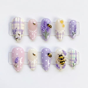 A set of ten almond-shaped press-on nails featuring lavender, white, and cream colors. Designs include lavender sprigs, small bees, white polka dots, and plaid patterns. One nail features a large, three-dimensional bee embellishment.
