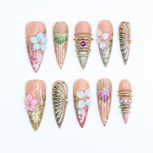 A set of ten stiletto-shaped press-on nails featuring a nude base with intricate gold filigree, 3D floral accents in blue, pink, and white, metallic gold rays, and embedded rhinestones, including heart motifs and stacked gold rings.