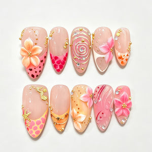 A set of ten almond-shaped press-on nails featuring a nude base with intricate pink and orange designs. Embellishments include three-dimensional floral accents, gold metallic seahorses, starfish, swirling lines, and small clear rhinestones.