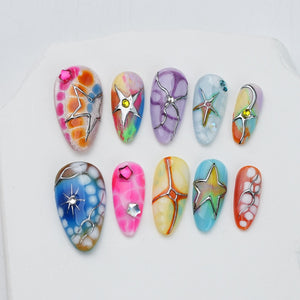 A set of ten almond-shaped press-on nails featuring diverse designs including multi-colored marble patterns, metallic silver star overlays, small embedded rhinestones, and intricate line work in shades of pink, blue, orange, and purple.