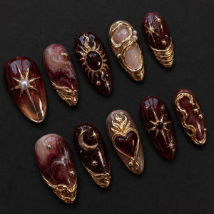 A set of ten almond-shaped press-on nails featuring a deep burgundy and cream marbled base. The designs include gold metallic accents, raised heart motifs, celestial sun and moon symbols, starbursts, and small white pearl beads.