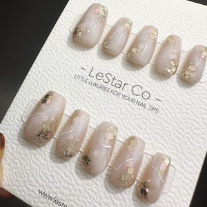 A set of ten press-on nails in a soft nude shade, featuring gold and pink glitter accents and thin white swirl lines. The nails are displayed on a white textured card labeled LeStar Co. - LITTLE LUXUR