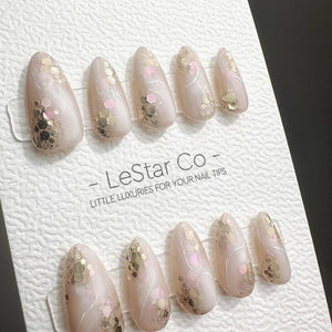 A set of ten almond-shaped press-on nails in a sheer nude base with gold and pink hexagonal glitter accents and thin white swirl lines. The nails are displayed on a white textured card labeled LeStar