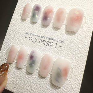 Ten press-on nails with a matte, translucent finish featuring soft watercolor-style swirls of peach, purple, and white with embedded iridescent flakes. The nails are displayed on a white card labeled