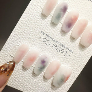 A set of ten rectangular press-on nail tips with a milky white base and soft watercolor-style smudges of purple, pink, and green. The tips are displayed on a white textured card labeled LeStar Co. - L