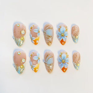 A set of ten almond-shaped press-on nails featuring a nude base with gradient orange and blue tips. The designs include raised 3D light blue flowers, gold metallic swirls, small white pearls, and iridescent rhinestones on each nail.