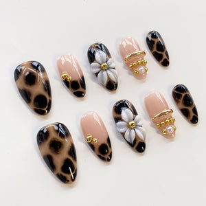 A set of ten press-on nails featuring a mix of designs including brown and black leopard print, solid nude bases with gold metallic beads, and nude nails adorned with white 3D flower petals and a central gold bead accent.