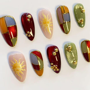 A set of ten almond-shaped press-on nails featuring a variety of designs including stained glass patterns in red, orange, and green with gold lines, solid deep red with gold beads, and pale pink with a yellow sun illustration.