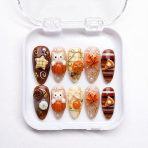 A set of ten almond-shaped press-on nails in a white plastic case. The designs feature autumn themes including 3D orange pumpkins, kittens, maple leaves, plaid patterns, brown buttons, and small slices of pumpkin pie with white cream.