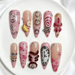 A set of ten almond-shaped press-on nails featuring a pink and nude color palette. Designs include silver swirls, a yellow banana charm, metallic rings, a star charm, a 3D flower, silver beads, pink stars, a monkey figure, and a clock.