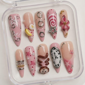A set of ten almond-shaped press-on nails in a clear plastic case. The designs feature pink and nude tones with various 3D embellishments including silver rings, a yellow banana, a star charm, a brown monkey, and a melting clock face.