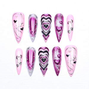 A set of ten almond-shaped press-on nails featuring a light pink base with metallic magenta and black accents. The designs include raised heart motifs, silver chrome line work, small silver studs, and dangling heart-shaped charms.