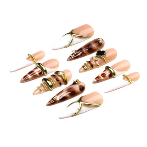 A set of ten stiletto-shaped press-on nails featuring a mix of nude, white, and brown cat-eye patterns. The nails are decorated with gold-toned metal rings, chains, small white pearls, and a black four-leaf clover charm accent.