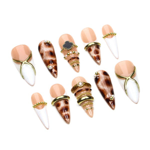 A set of ten press-on nails with a pointed shape. The designs include nude and white French tips with gold cross accents, brown leopard print patterns, and various embellishments such as gold rings, small pearls, and a black clover.