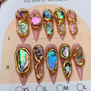 A set of ten stiletto-shaped press-on nails with a light pink base. Each nail features irregular, iridescent abalone shell fragments encased in raised, textured gold-colored metal borders. The nails are arranged on a display card.