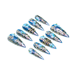A set of ten stiletto-shaped press-on nails featuring an iridescent blue finish, raised silver filigree scrollwork, small light blue floral accents, and select nails adorned with horizontal bands of embedded clear rhinestones and gold beads.